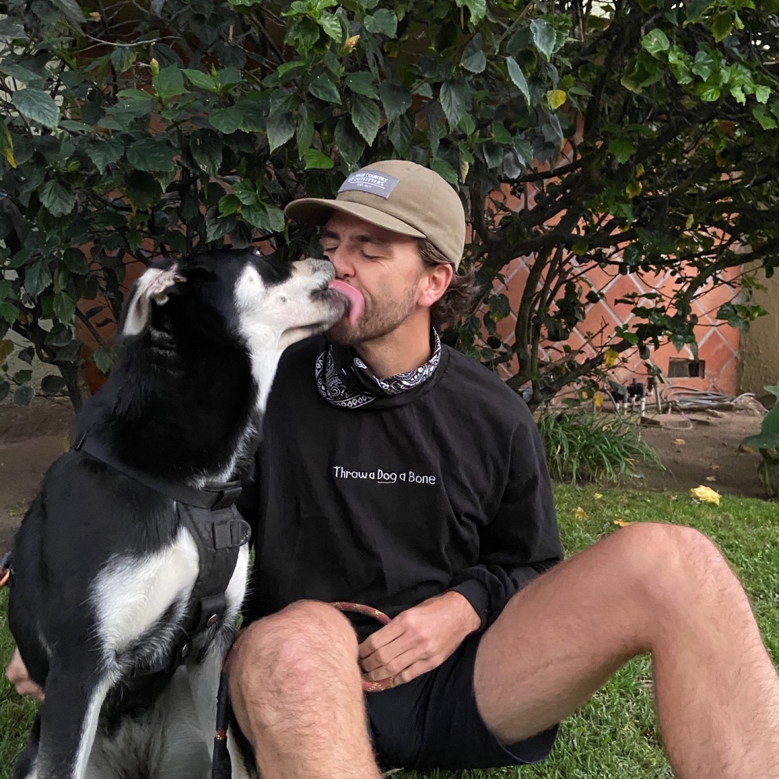 A man wearing a black sweatshirt embroidered with the phrase 'Throw a dog a bone' is licked by a black and white dog